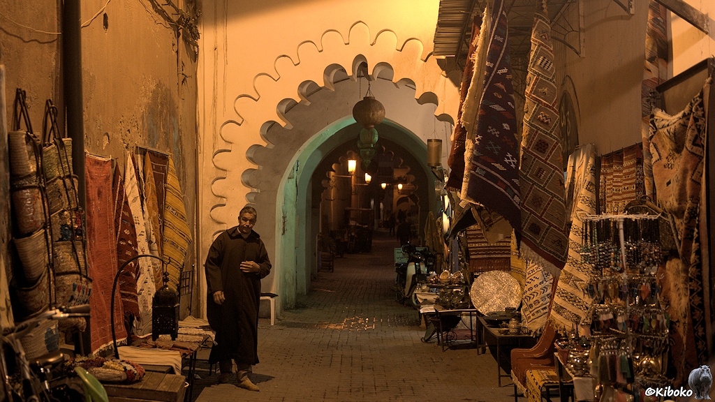 Bis spät in die Nacht präsentiert ein Teppichhändler seine Waren in einer Gasse vor einem Bogen mit gezackte Wänden.