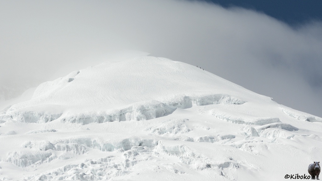 Das Bild zeigt die Schneekuppe von einem Vulkan. Hohe Stufen durchziehen den Gletscher. Am rechten Hang besteigen drei Menschen den Vulkan. Die Spitze und die linke Seite werden von einem Wolkenschleier verdeckt.