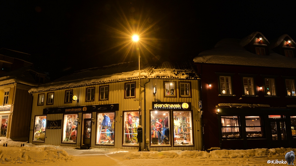 Das Bild zeigt ein Eckhaus. Durch die großen Schaufenster mit Sportbekleidung ist der beleuchtete Laden sichtbar. Schnee türmt sich vor dem Haus und auf dem Dach.