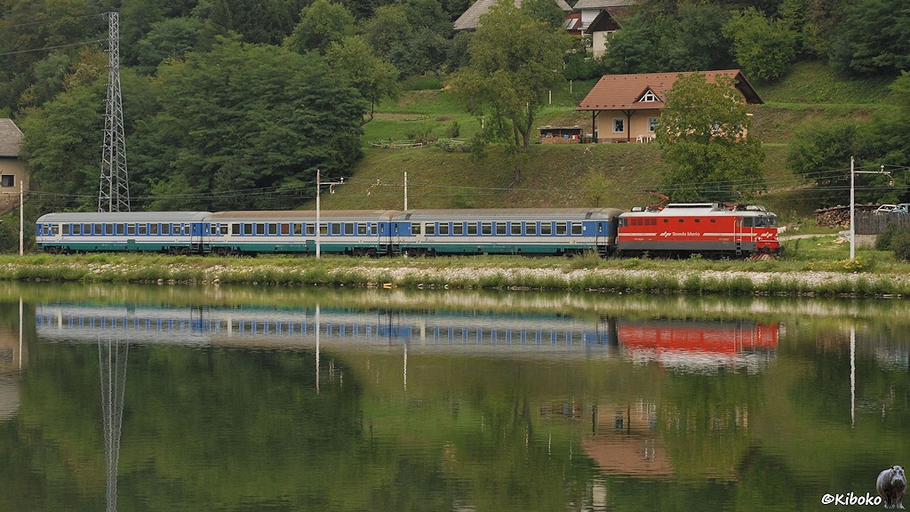 Eine rote Elektrolok mit drei blau-grau-grünen Personenwagen spiegelt sich in einem Fluss.