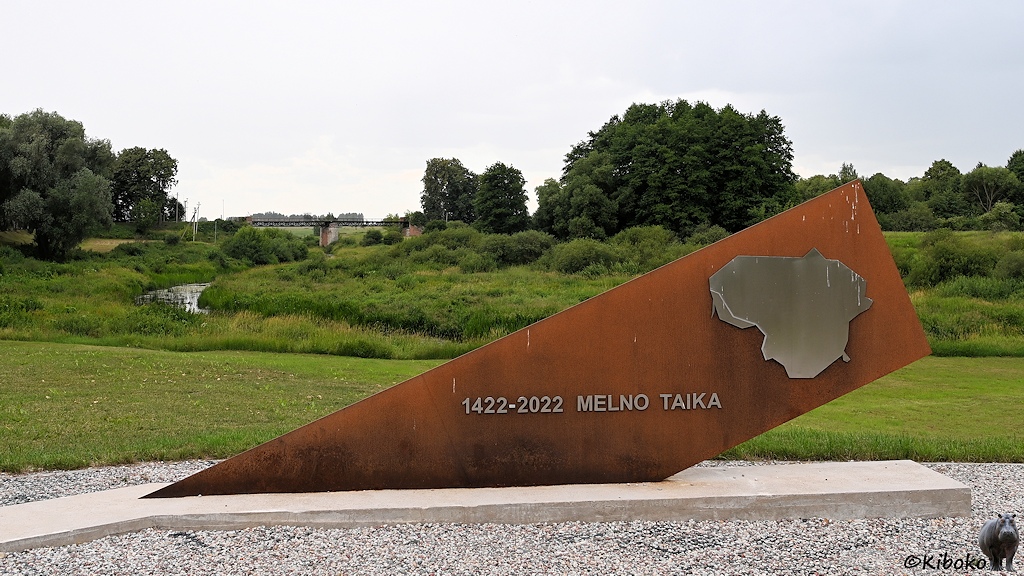 Das Bild zeigt ein rostfarbenes Denkmal aus einer schräg nach rechts geneigten Stahlplatte mit der Aufschrift 1422-2022 MELNO TAIKA. Dahinter ist eine Wiesenlandschaft durch die ein kleiner Fluss fliesst. Im Hintergrund führt eine Unterflur Gitterbrücke über diesen Fluss.
