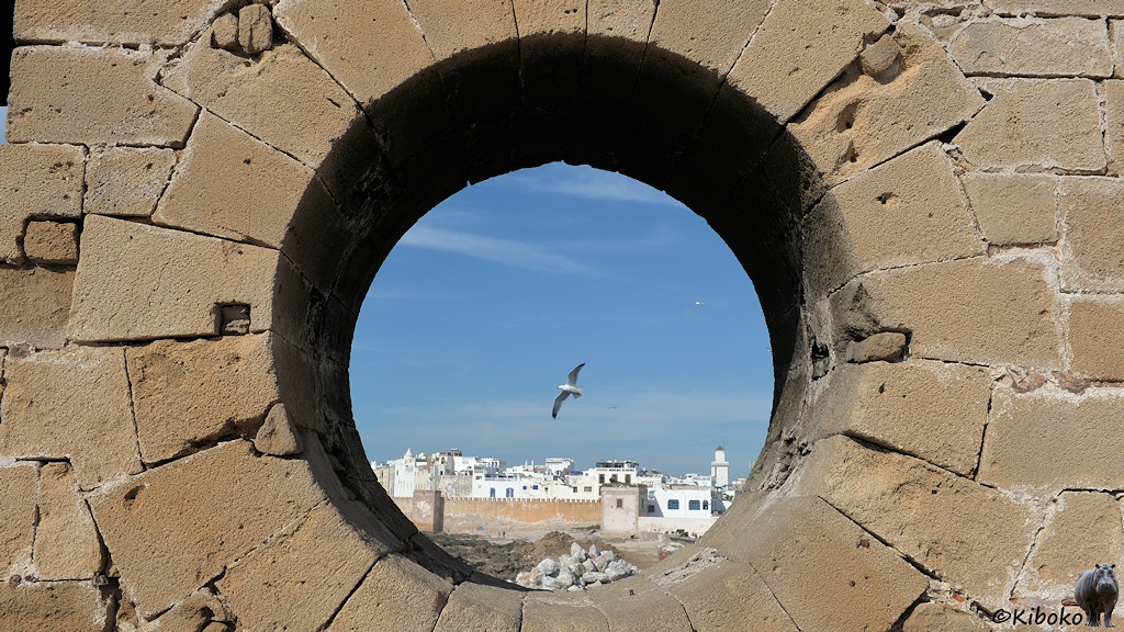 Das Bild zeigt ein rundes Loch in einer braunen Sandsteinmauer. Der Durchblick zeigt weiße Häuser unter blauem Himmel und eine fliegende Möwe.