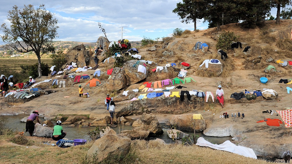 Frauen waschen Wäsche in einem kleinen Fluß. Bunte Wäschestücke trocknen auf den umliegenden Felsen.